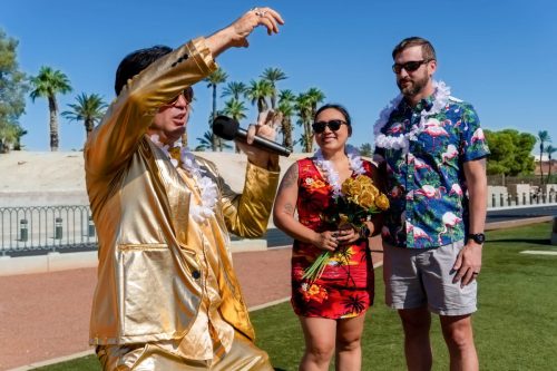 Gold-suited Elvis officiates a wedding for a bride and groom holding a bouquet at the Las Vegas sign.