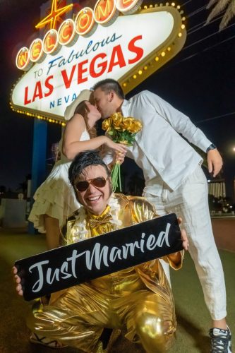 Happy couple wearing sunglasses pose with an Elvis impersonator at the Welcome to Fabulous Las Vegas sign.