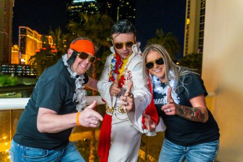 Bride and groom pose with a white and red jumpsuit-clad Elvis impersonator at a picnic table during their Las Vegas sign wedding.