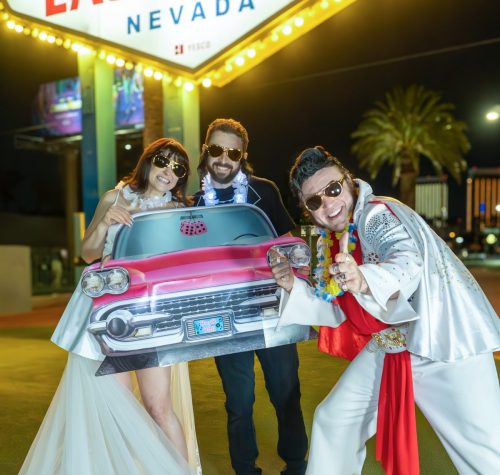 Bride and groom pose with a 'Las Vegas' license plate and a white-jumpsuit Elvis impersonator at the Welcome to Fabulous Las Vegas sign.