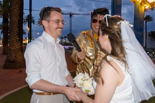 Bride holds her bouquet while embracing the groom and smiling with a gold-suited Elvis impersonator during their Las Vegas sign wedding.