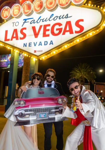 Couple wearing sunglasses smile under the Nevada Las Vegas sign during their Elvis-themed wedding.