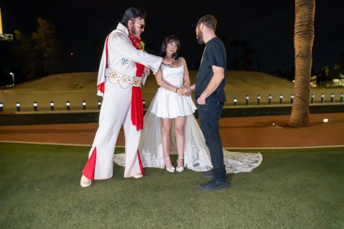 Elvis impersonator in a white and red suit poses with bride and groom on a grassy lawn at night during their Las Vegas wedding.
