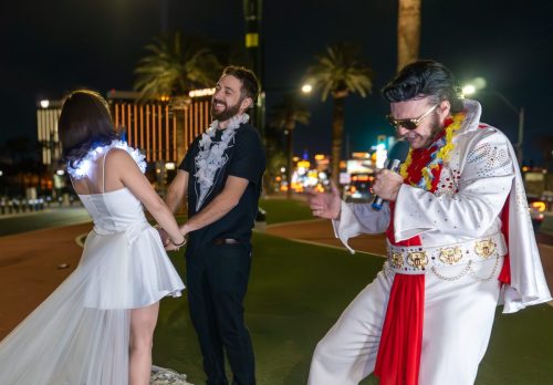 Bride twirls with Elvis impersonator in a white and red jumpsuit while the groom cheers during their Las Vegas sign wedding.